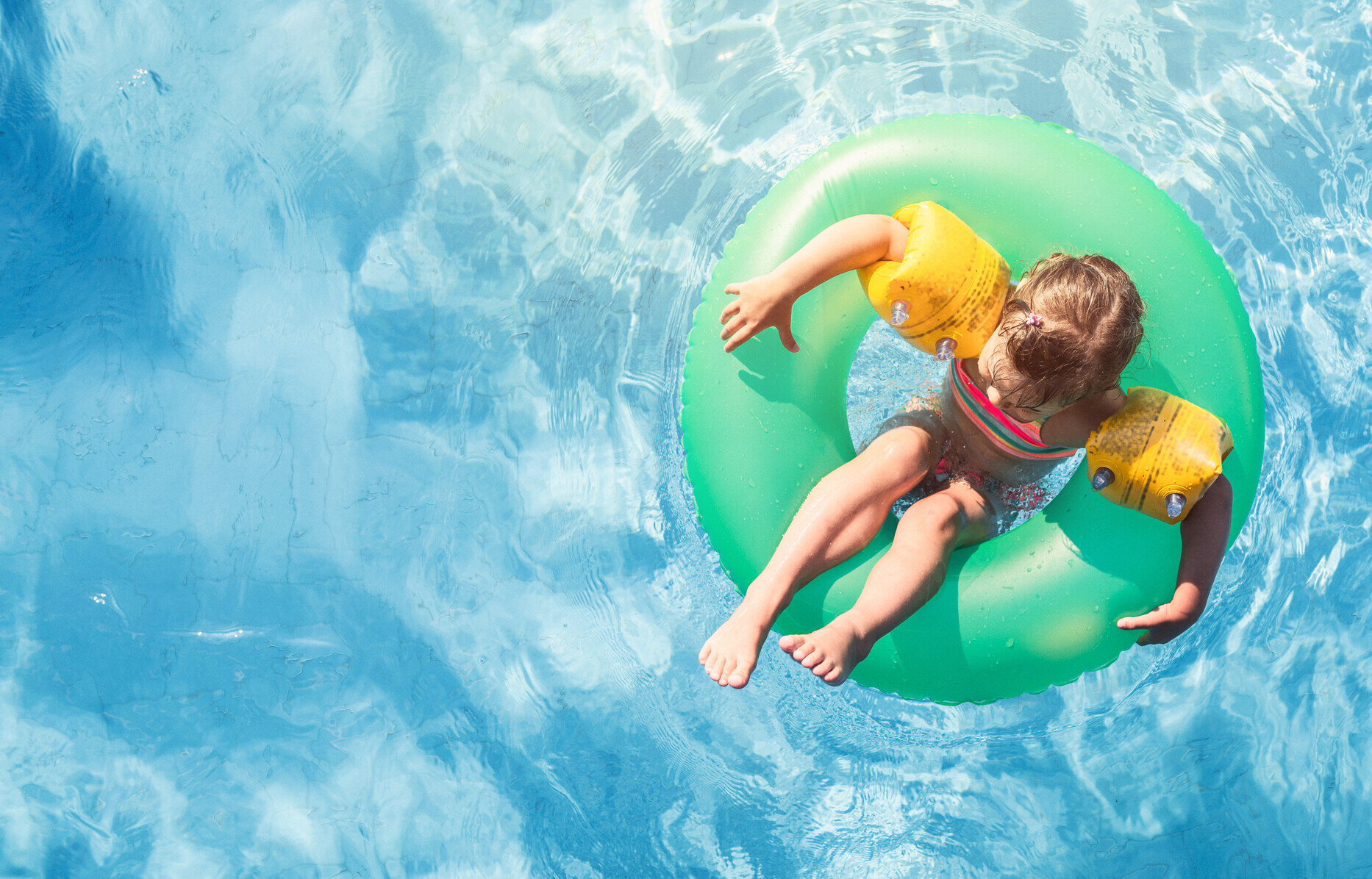 A child wearing yellow floaties and sitting on a green pool float in a swimming pool.
