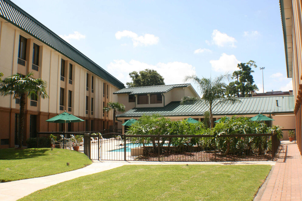 A tan and brown hotel building with a green metal roof, a fenced pool area, and palm trees on a green lawn.